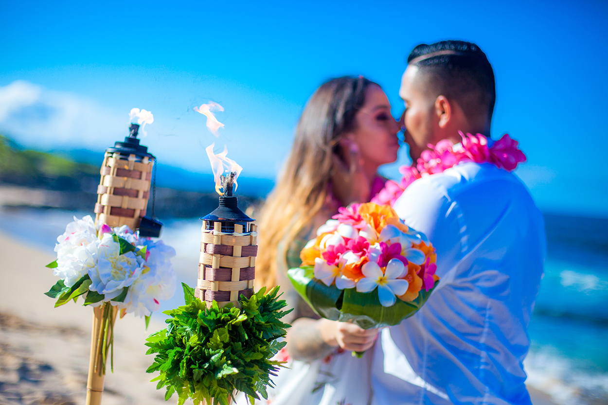 Couple about to kiss beside lit tiki torches, with the bride holding a rainbow rose bouquet